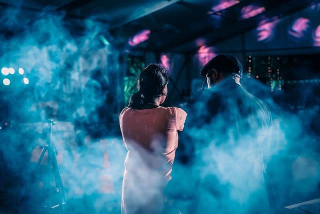 A woman and a man seen from behind, standing in blue haze under stage lights