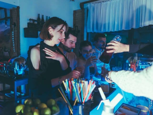 Friends at a blue-lit bar, one checking her phone while a bartender pours a drink