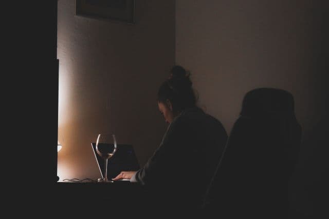 A woman typing on a laptop at a dark desk, lit by a small lamp, with a glass of wine beside her