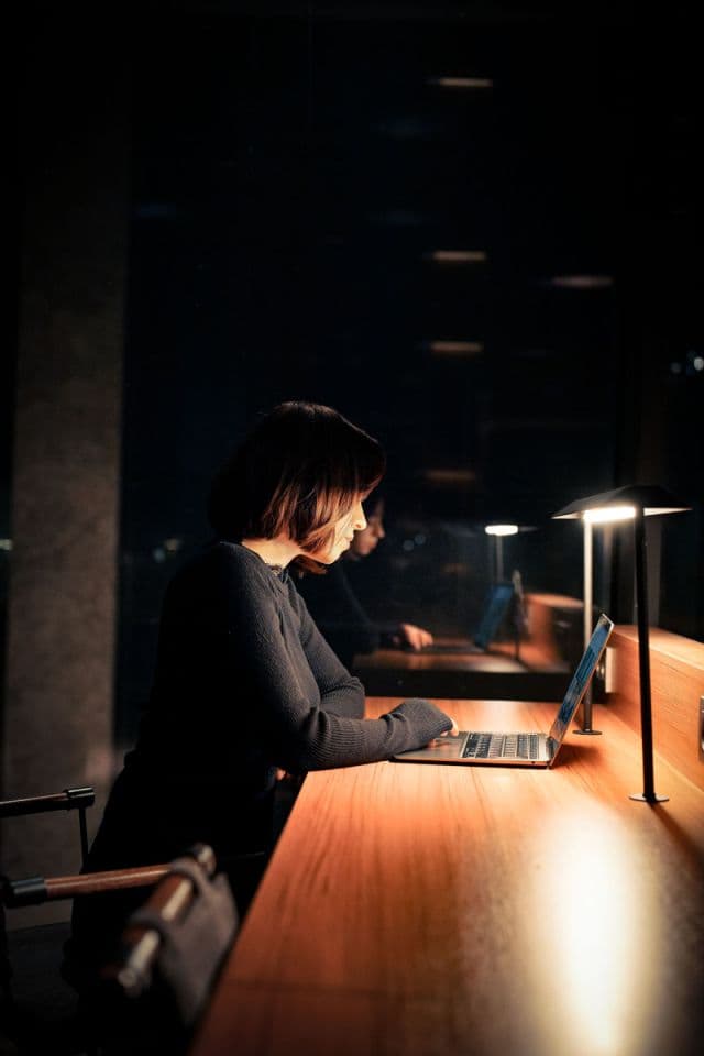 A woman working late at a laptop under a desk lamp in a dim office