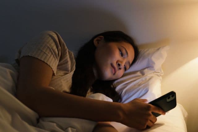 A woman lying in bed at night, reading a message on her phone by warm lamplight