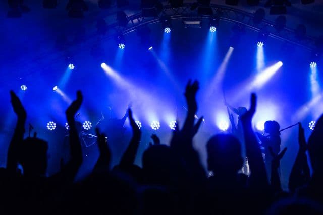 Silhouetted crowd with hands raised toward a band on a stage lit by bright blue spotlights