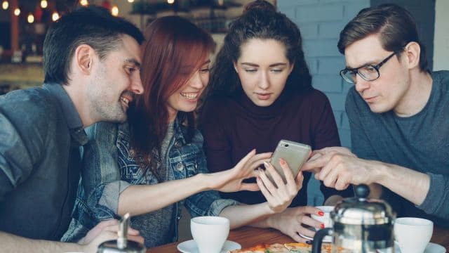 Four friends at a cafe table leaning in to look at one phone together