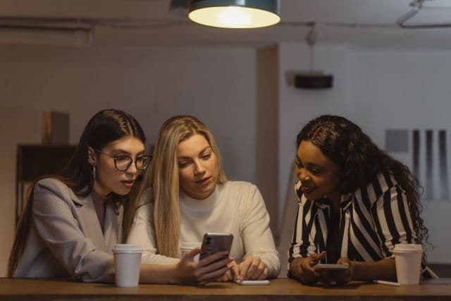 Three friends at a table leaning in to look at a smartphone together