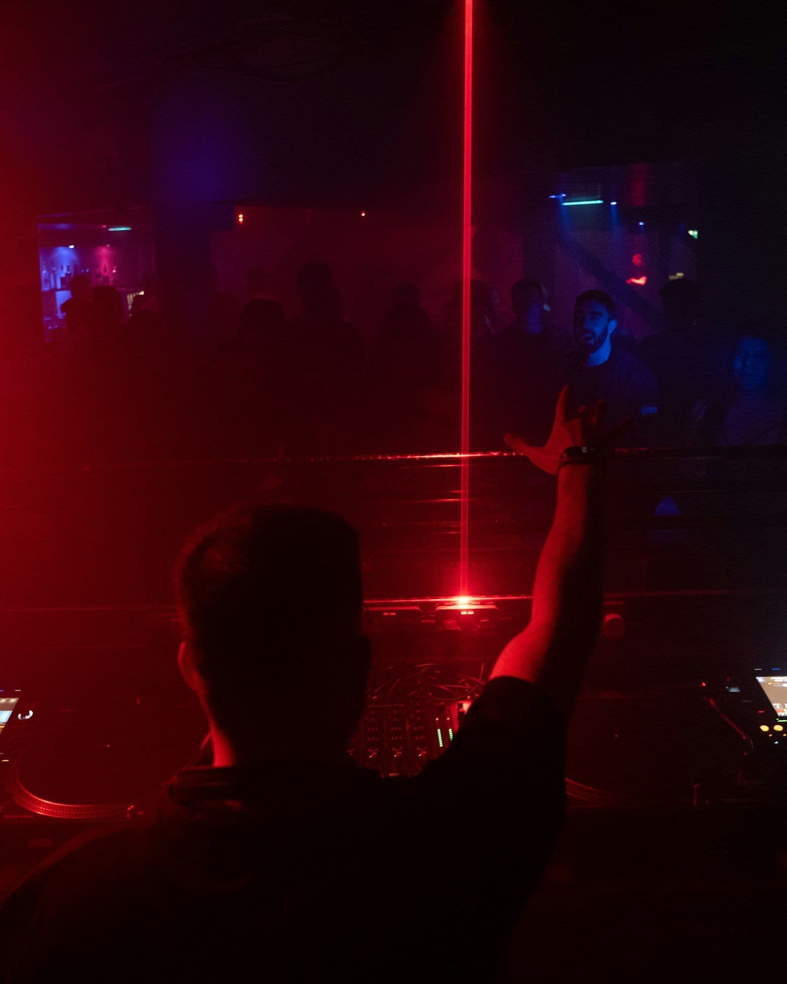 A DJ seen from behind raising a hand toward a crowd under a red laser