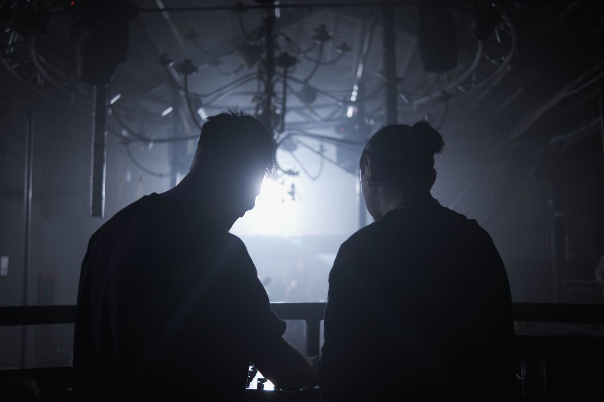 Two people in silhouette working side by side at a DJ booth in a smoky club