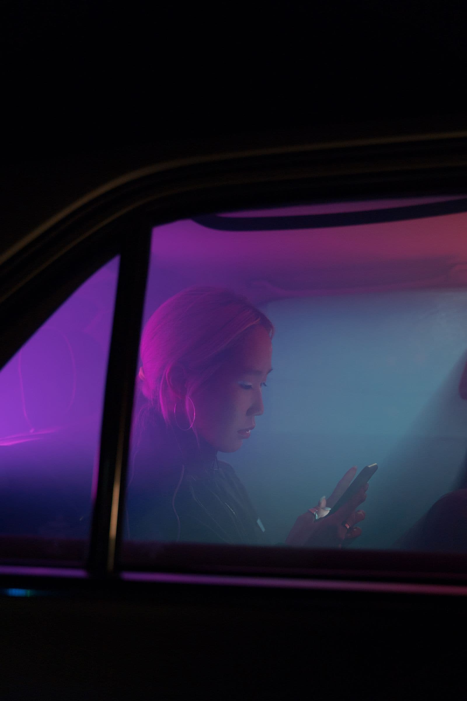 A woman lit by pink neon light checking her phone in a car at night