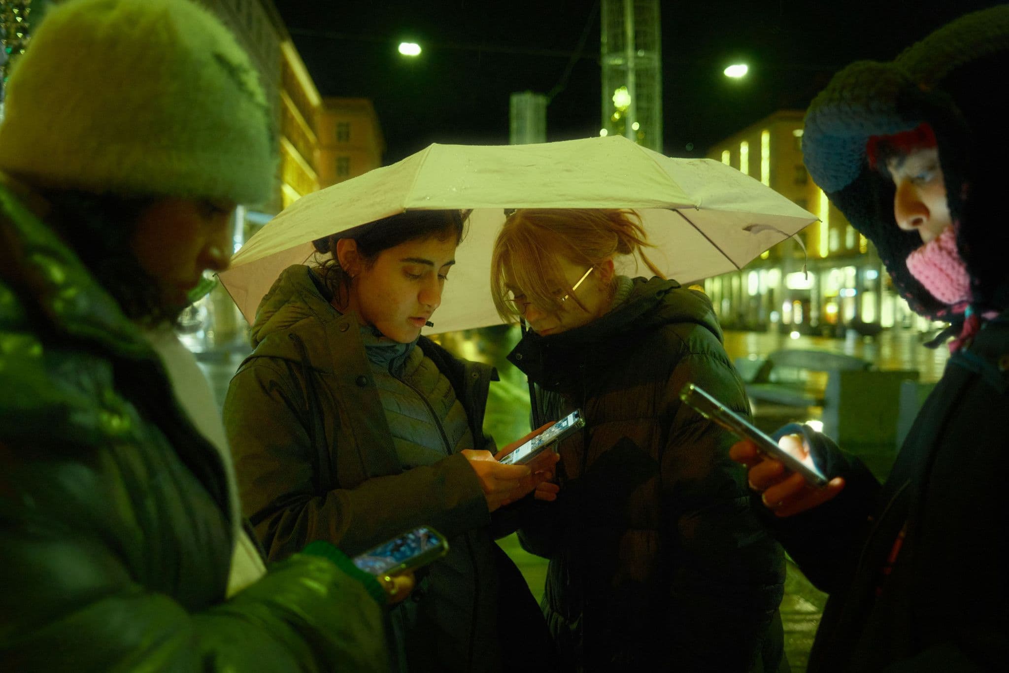 Friends huddled under an umbrella on a rainy street at night, checking their phones