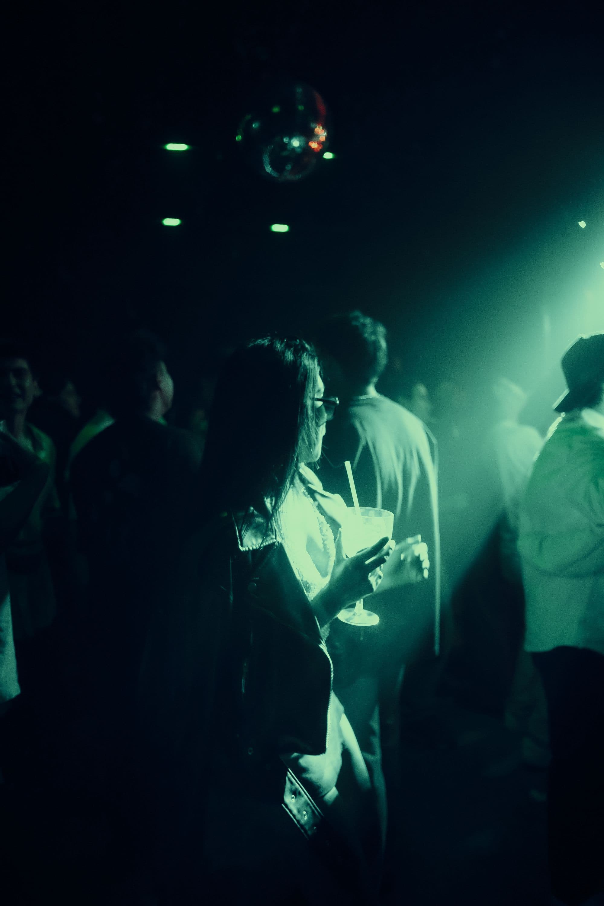 Small late-night crowd on a dark dance floor under green light and a mirror ball, one person holding a drink