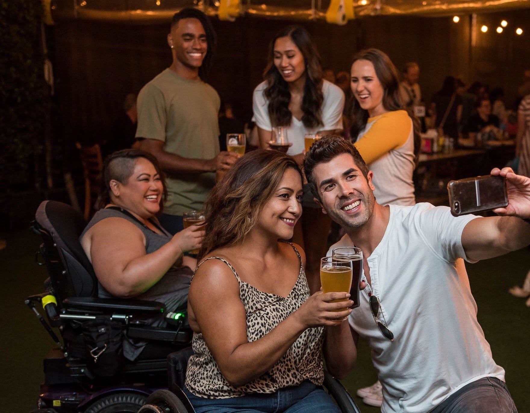 A diverse group of friends at an evening party under string lights, smiling and raising drinks as one of them takes a group selfie