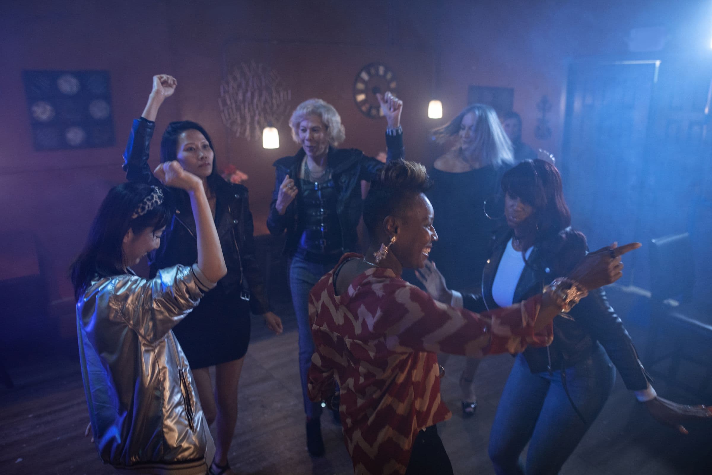 A diverse group of women dancing at a club after dark, one in a shiny metallic silver jacket
