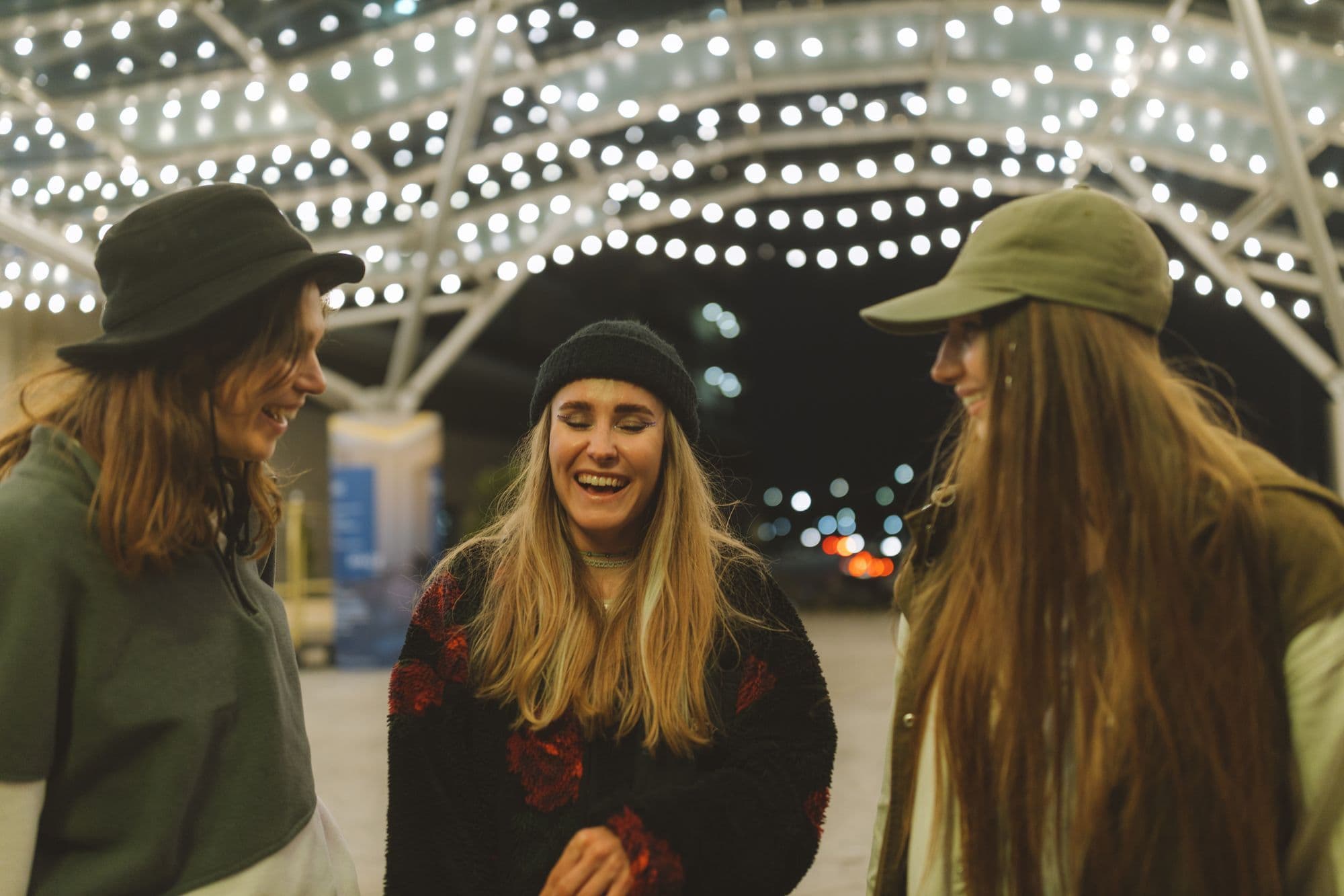 Three friends laughing together under strings of white lights on a night out