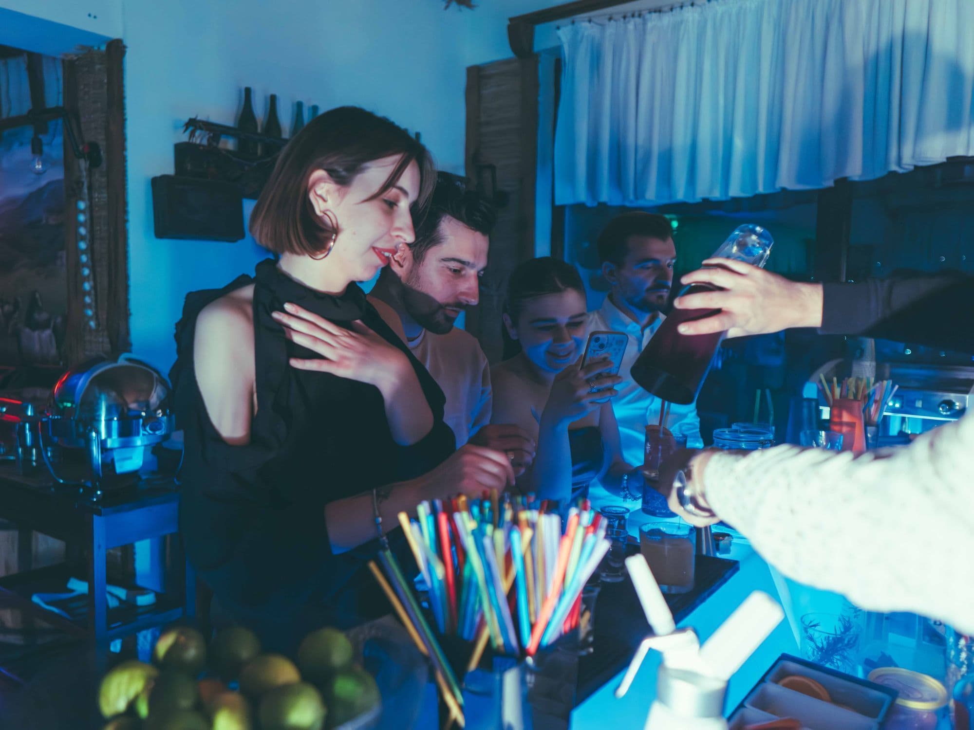 Friends at a blue-lit bar, one checking her phone while a bartender pours a drink