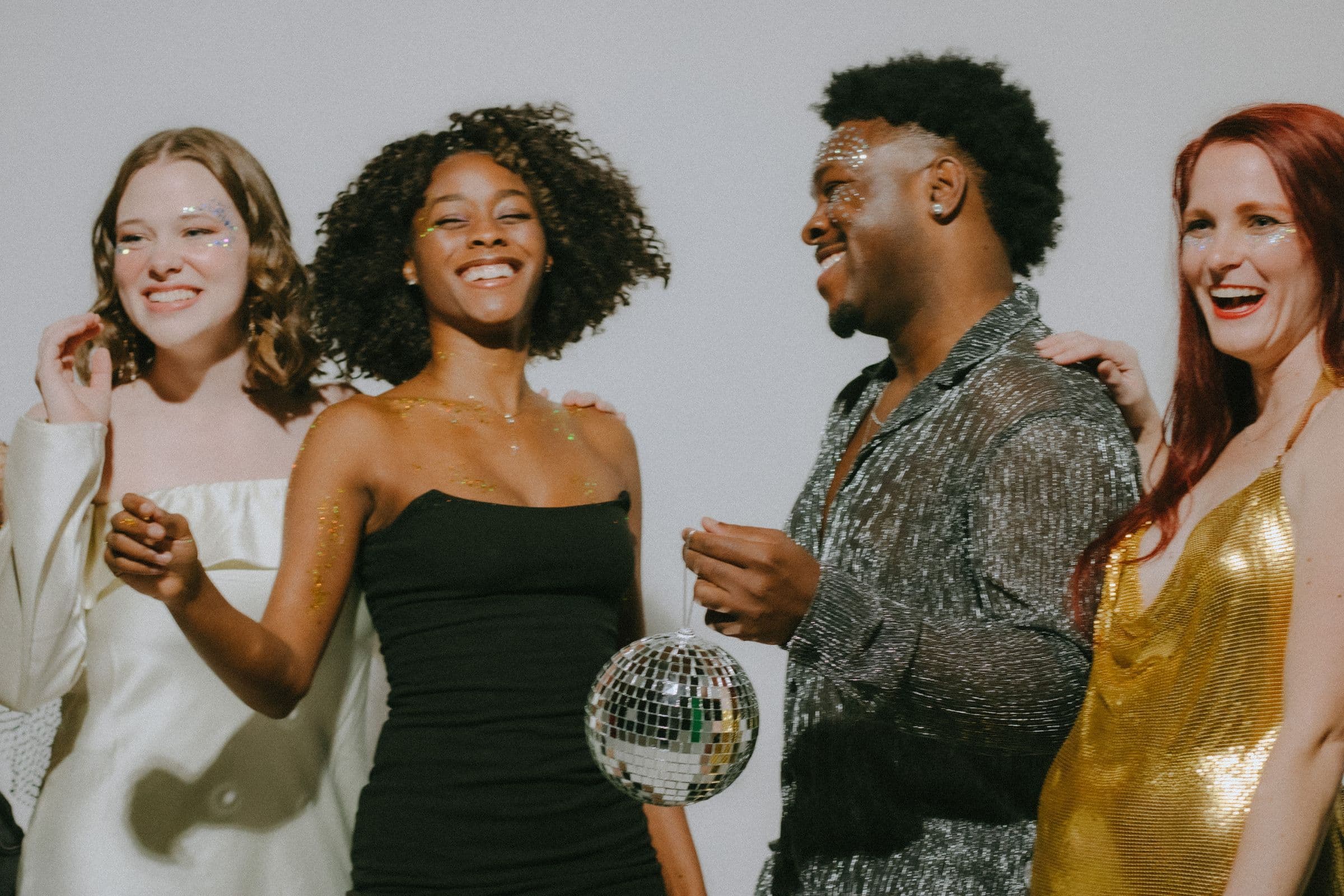 Four diverse friends in glitter makeup and metallic party outfits laughing together, one holding a small disco ball
