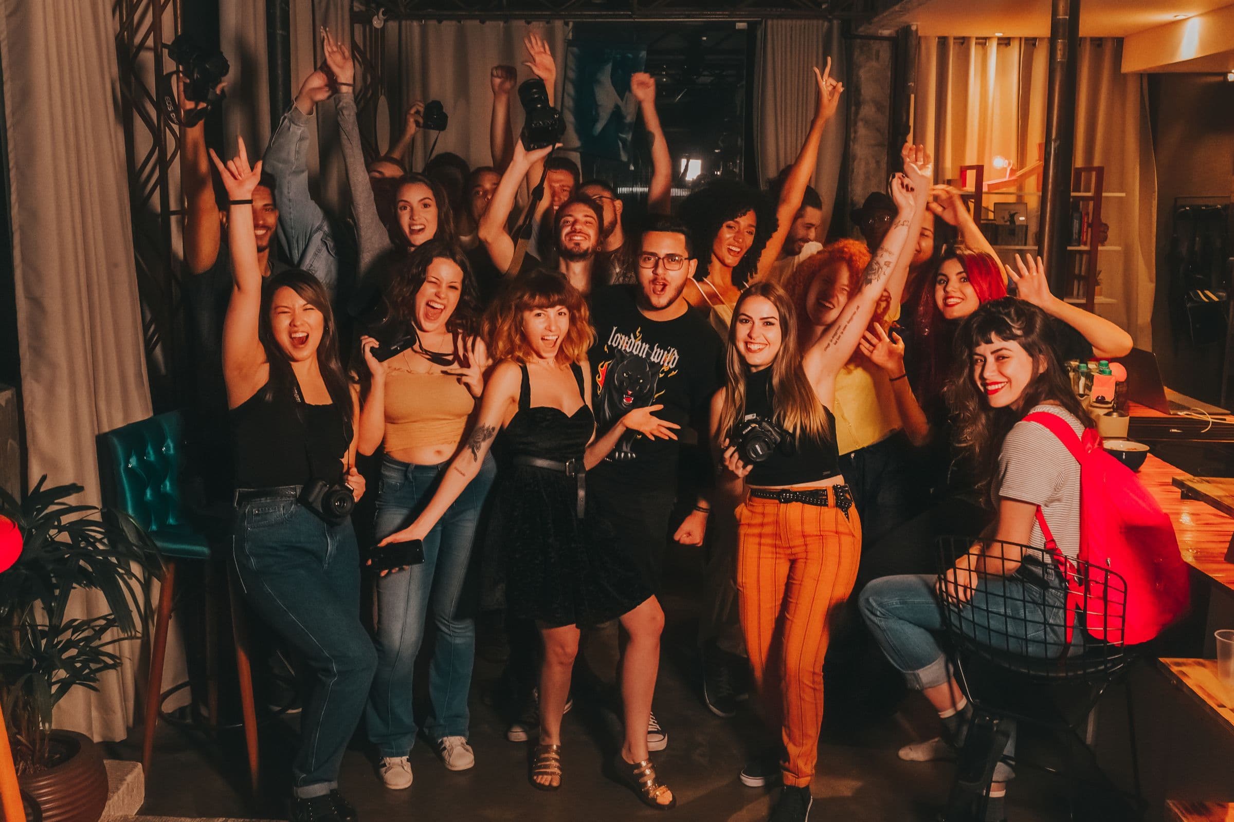 A diverse crowd of friends cheering with their hands up at a party in a warmly lit bar after dark