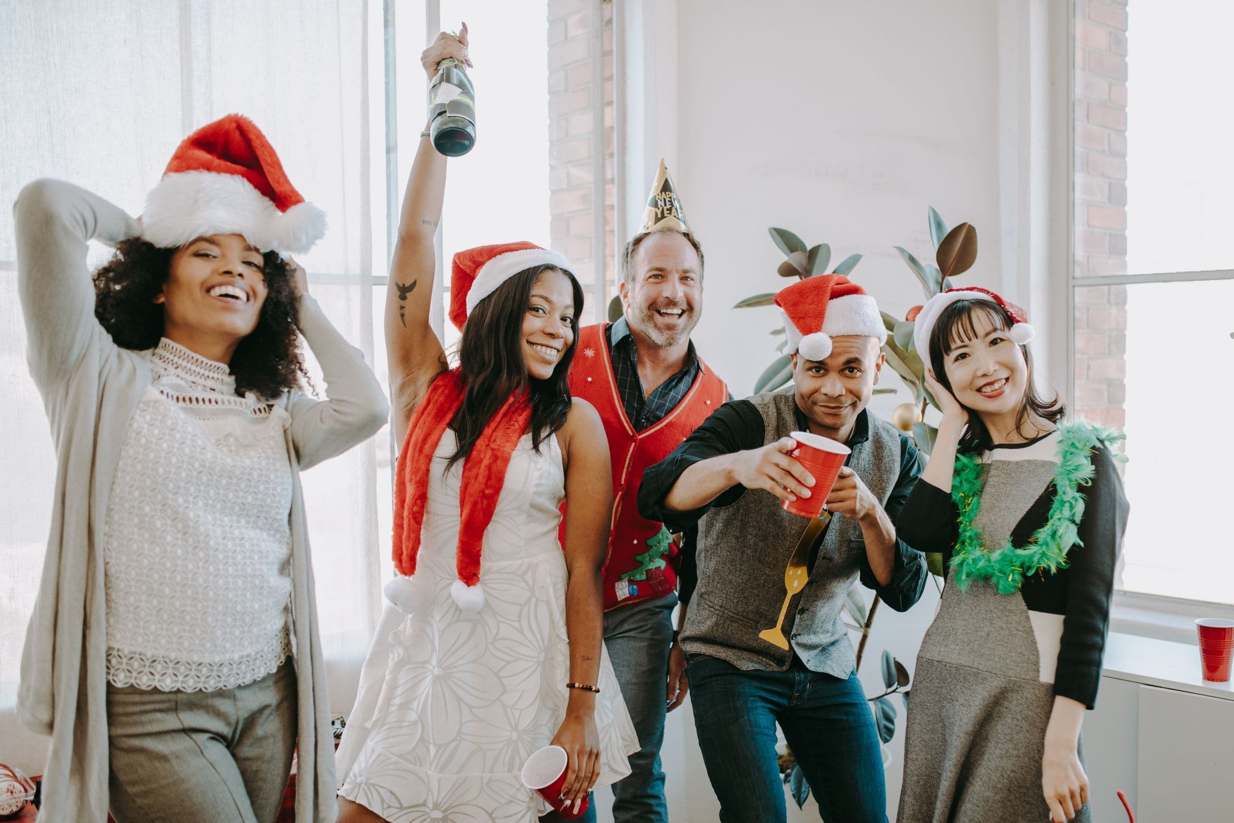 A diverse group of friends in Santa hats and festive outfits laughing and raising drinks at a holiday party