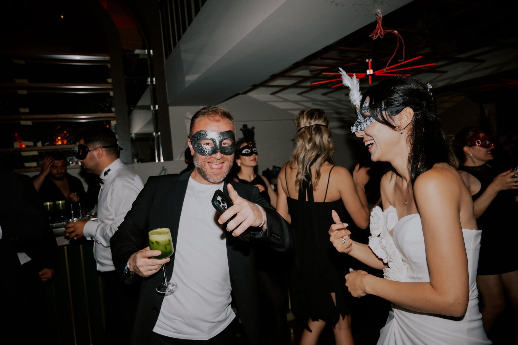 Guests in black and silver masquerade masks laughing and dancing at an indoor party, one holding a cocktail