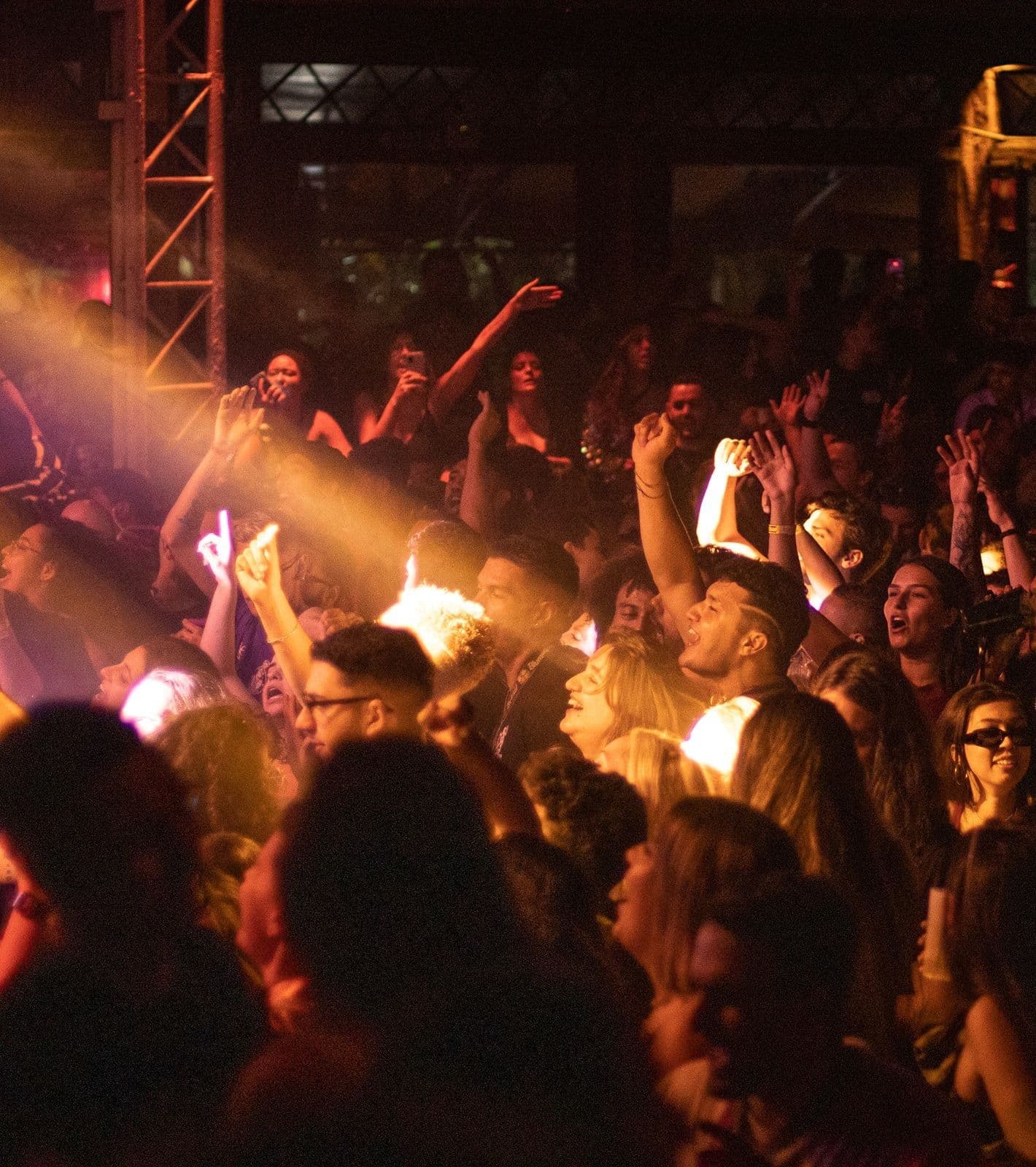 A diverse crowd raises their hands and sings along in a dark venue lit by a warm stage light