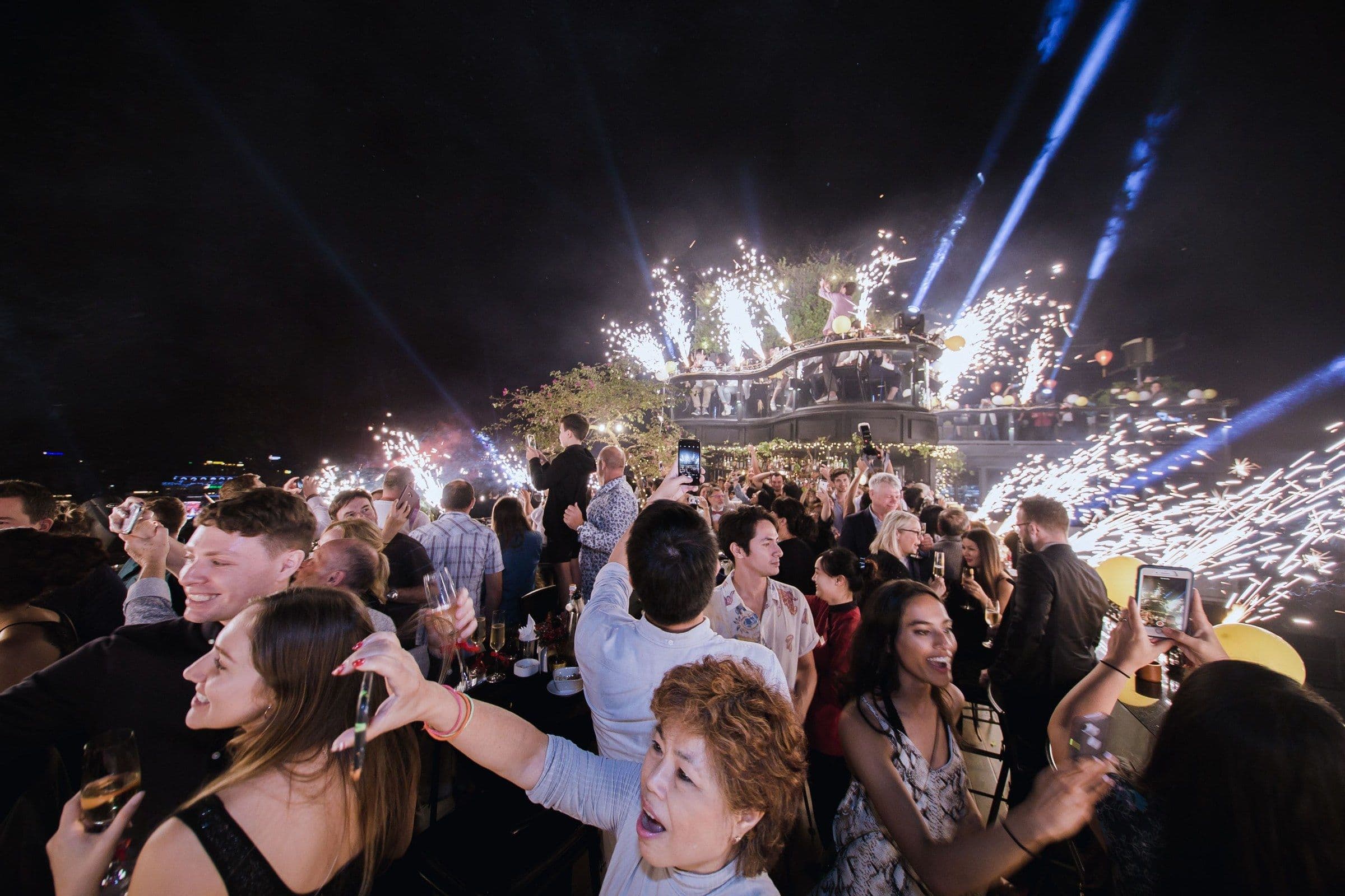 A diverse crowd cheers with drinks raised on a rooftop after dark as sparklers light up the stage behind them