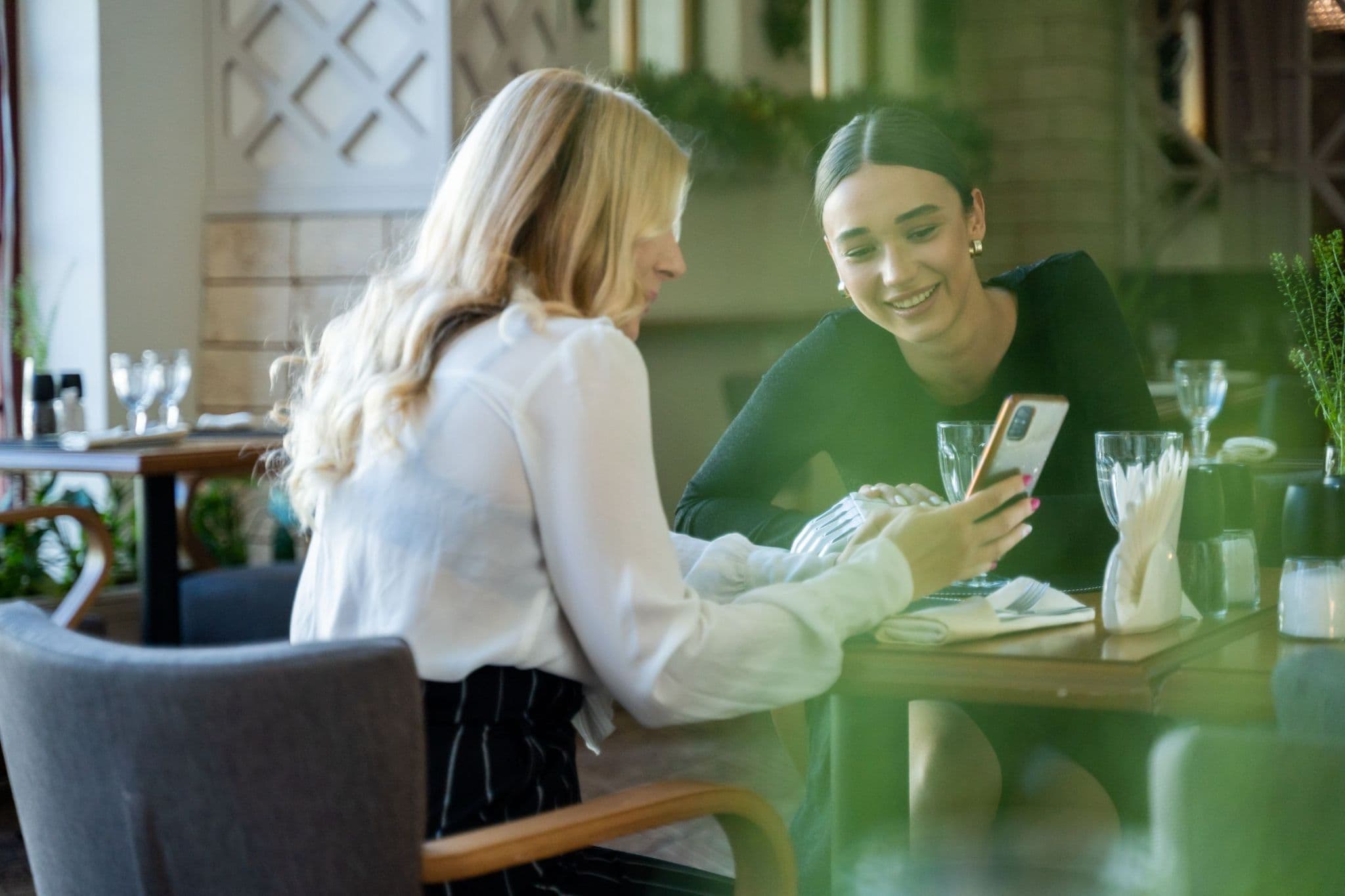 Two women at a restaurant table, smiling as they look at a phone together
