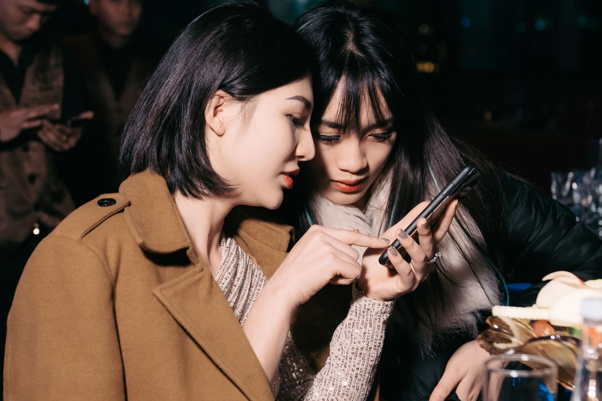 Two friends at a dim bar leaning in together to look at one phone