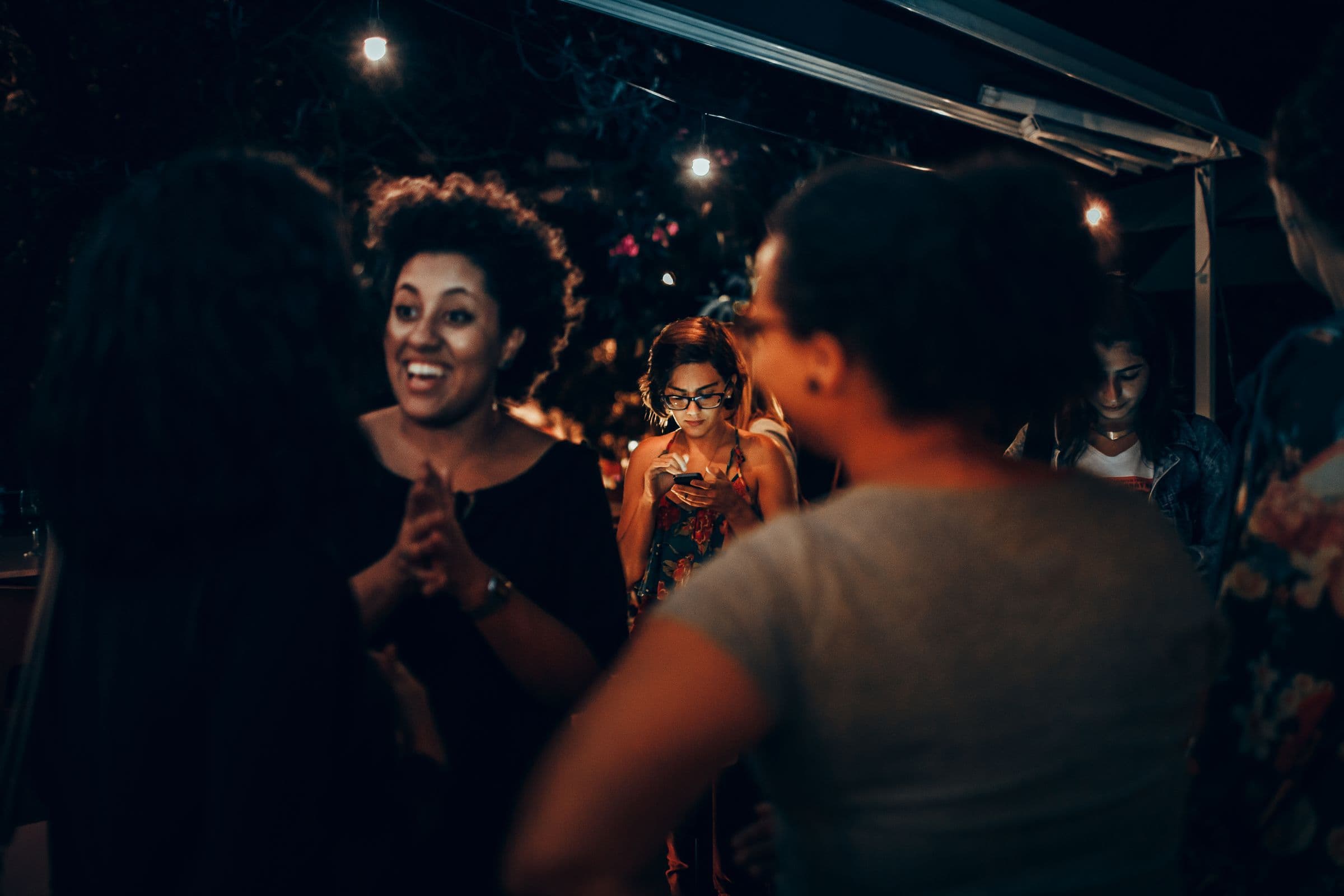 Friends of different backgrounds laughing and talking at an evening party under string lights, one checking her phone