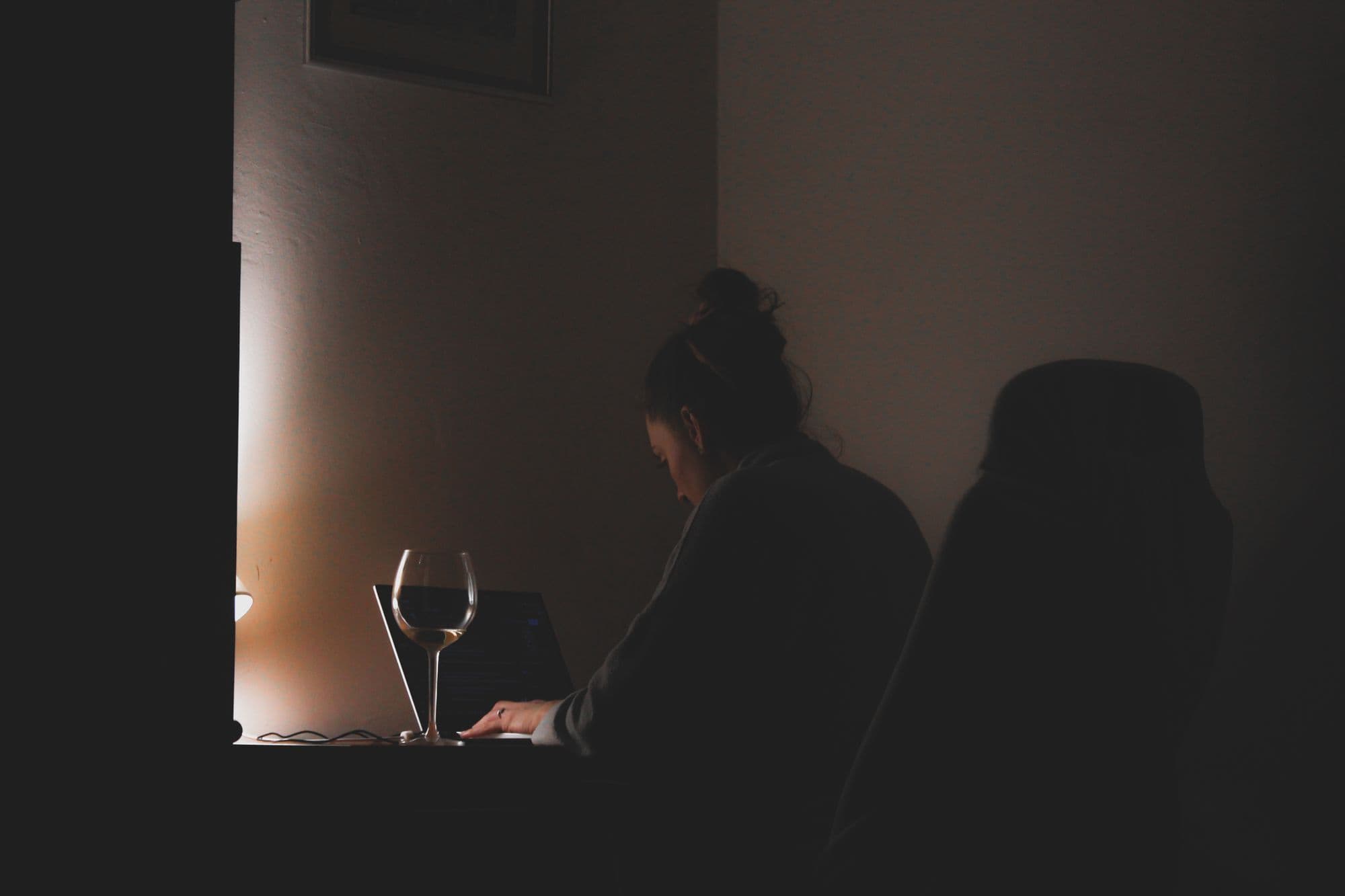 A woman typing on a laptop at a dark desk, lit by a small lamp, with a glass of wine beside her