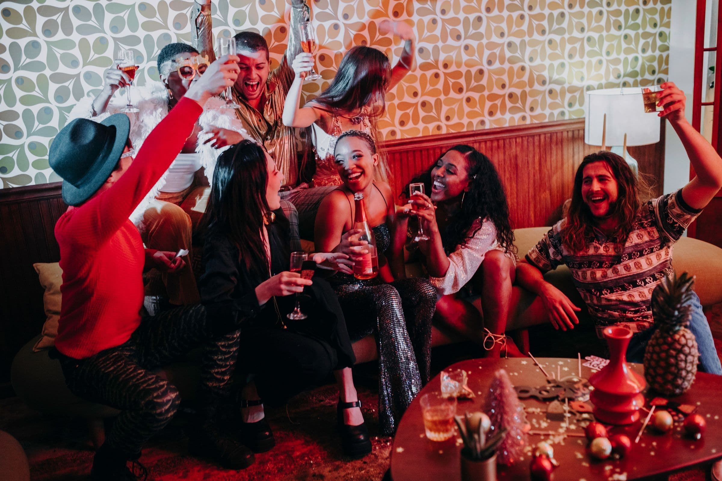A diverse group of friends in party outfits cheering and raising glasses on a couch at an evening holiday party, with ornaments on the table