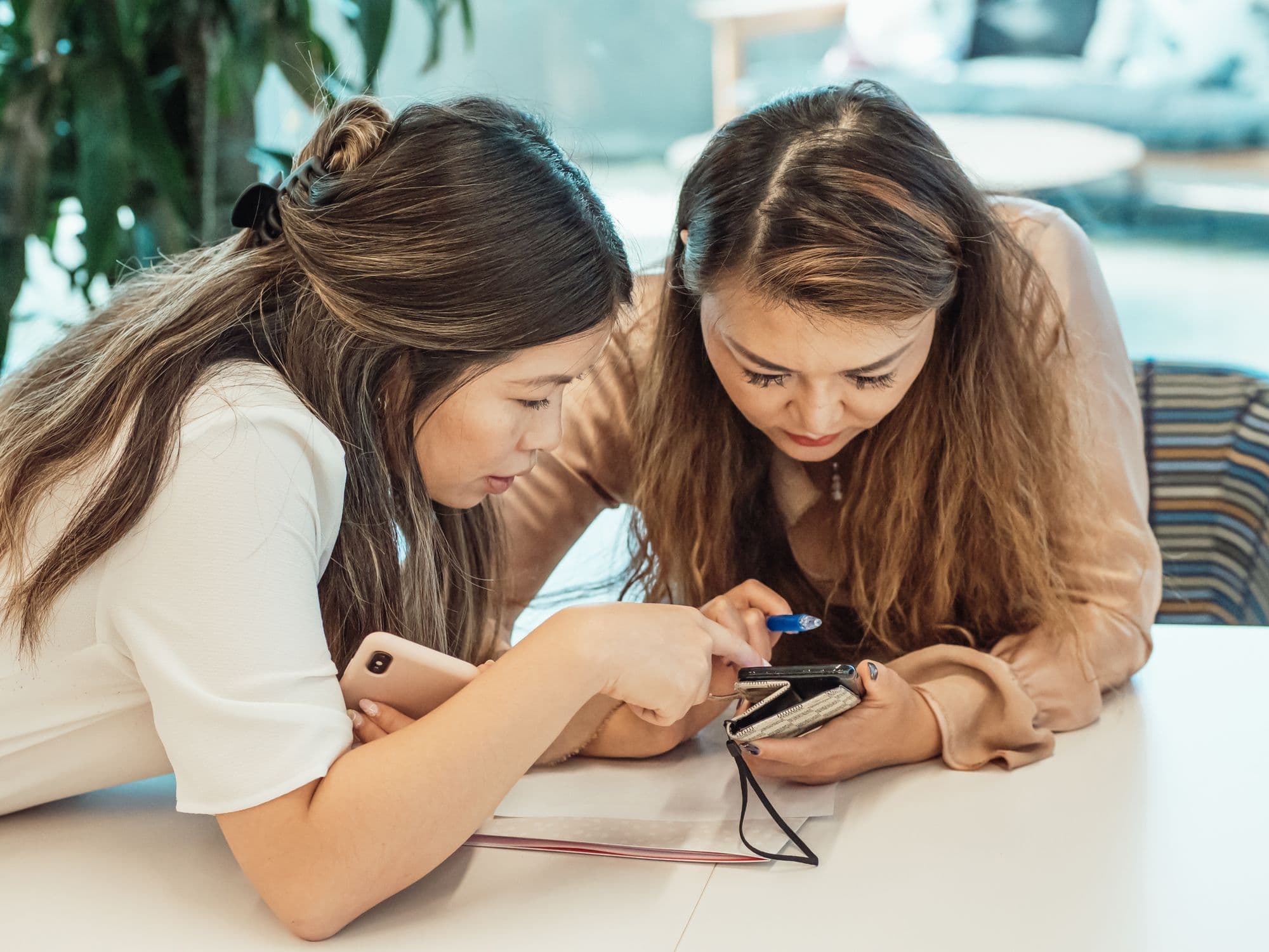 Two women leaning over a table and looking at one phone together