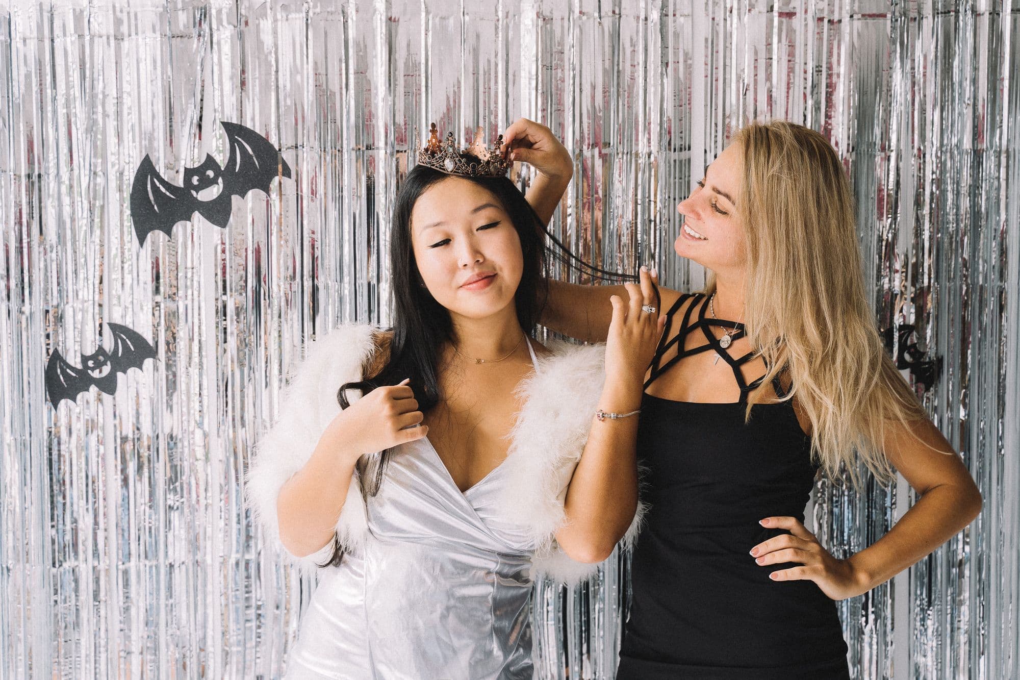 Two women in Halloween costumes, one adjusting the other's dark crown, in front of a silver foil curtain decorated with paper bats