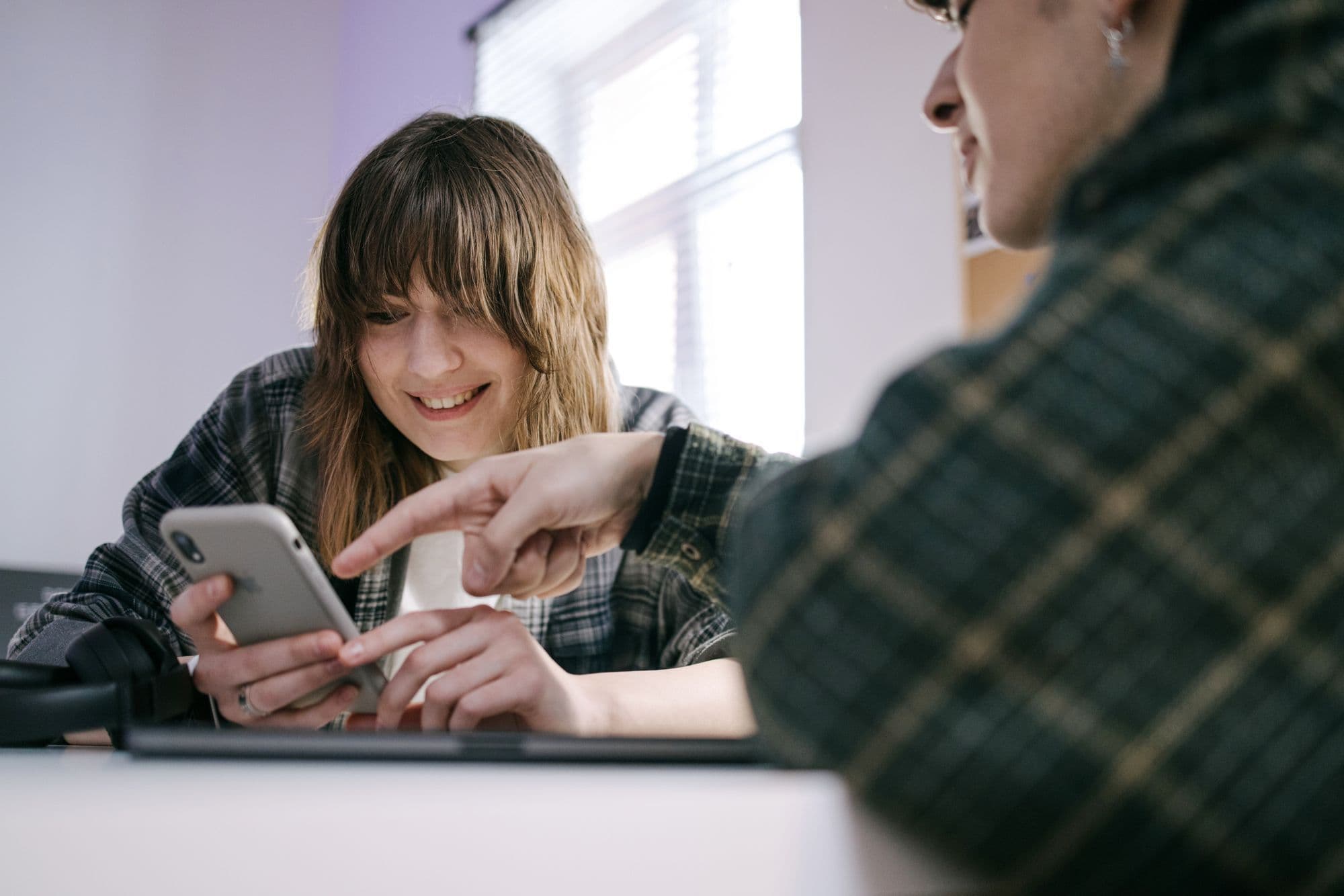 One person points at a phone while the other holds it and smiles, both sitting at a table
