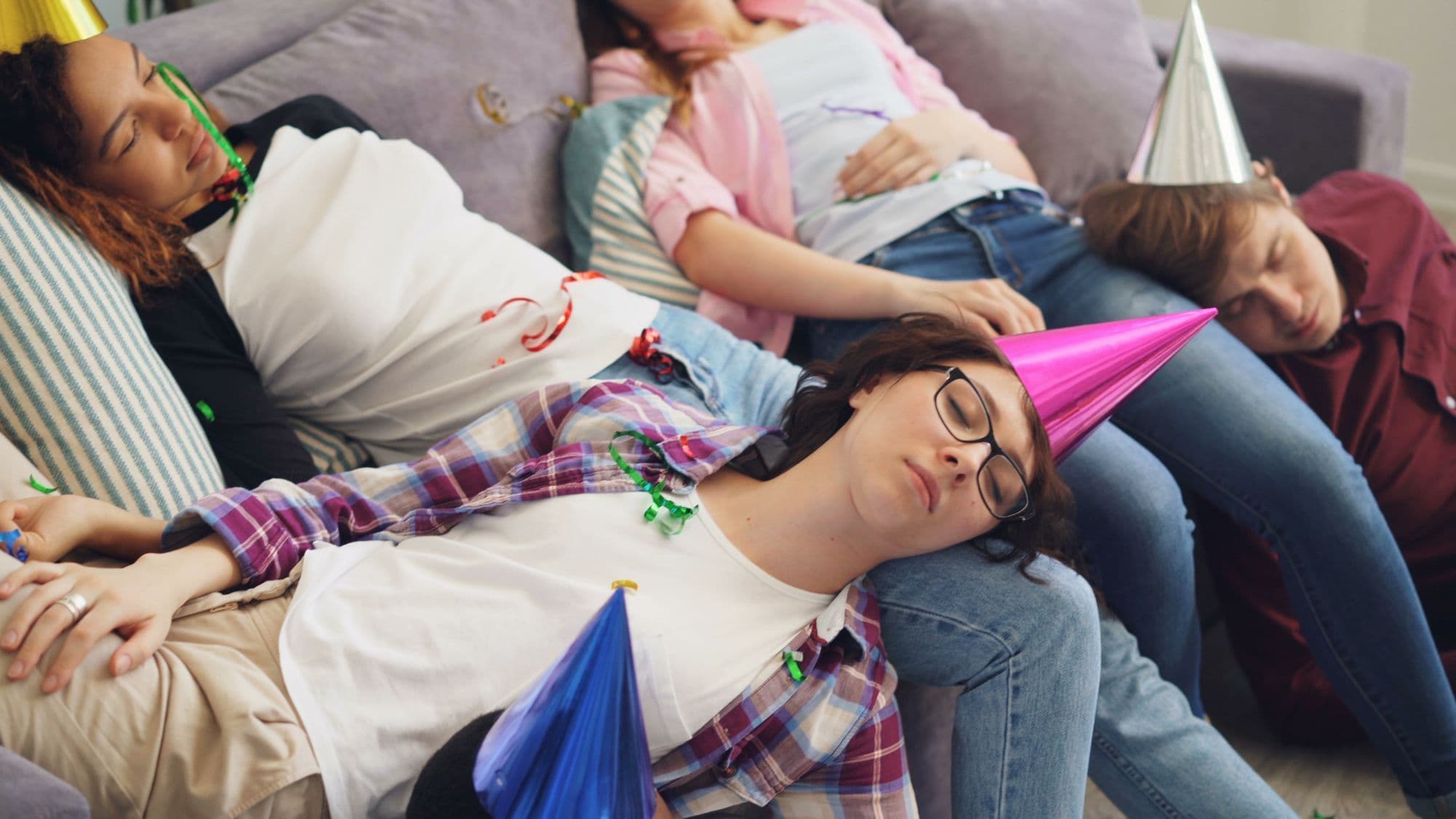 Four friends asleep on a couch in party hats, streamers still on them