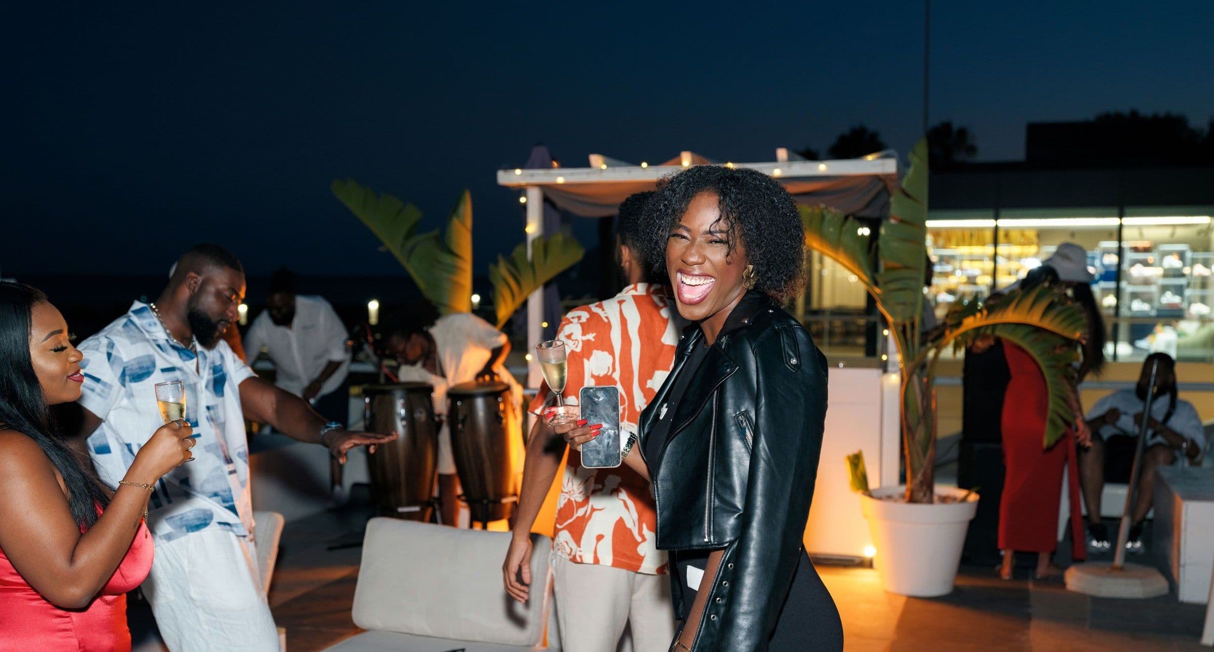 Guests laugh and dance on a palm-lined rooftop lounge after dark, with drums set up behind them