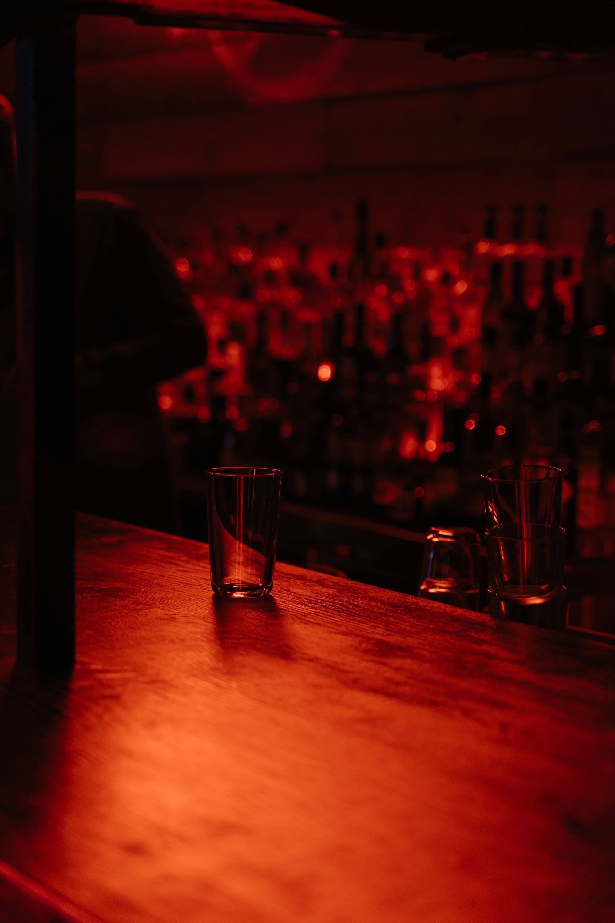 Empty glass on a wooden bar counter under deep red light with bottles blurred behind