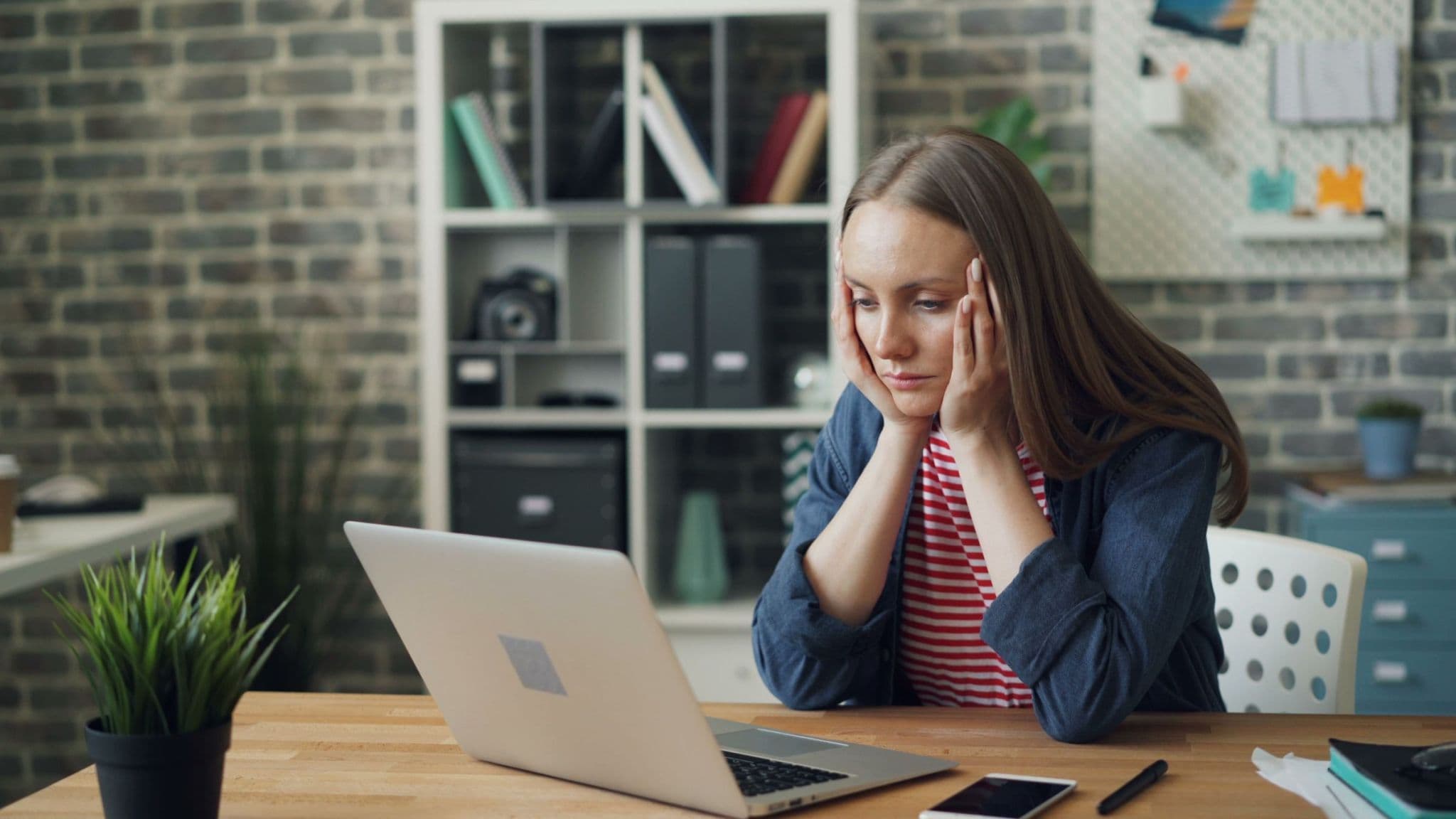 A woman at a desk with her head in her hands, staring at her laptop in an office with a brick wall