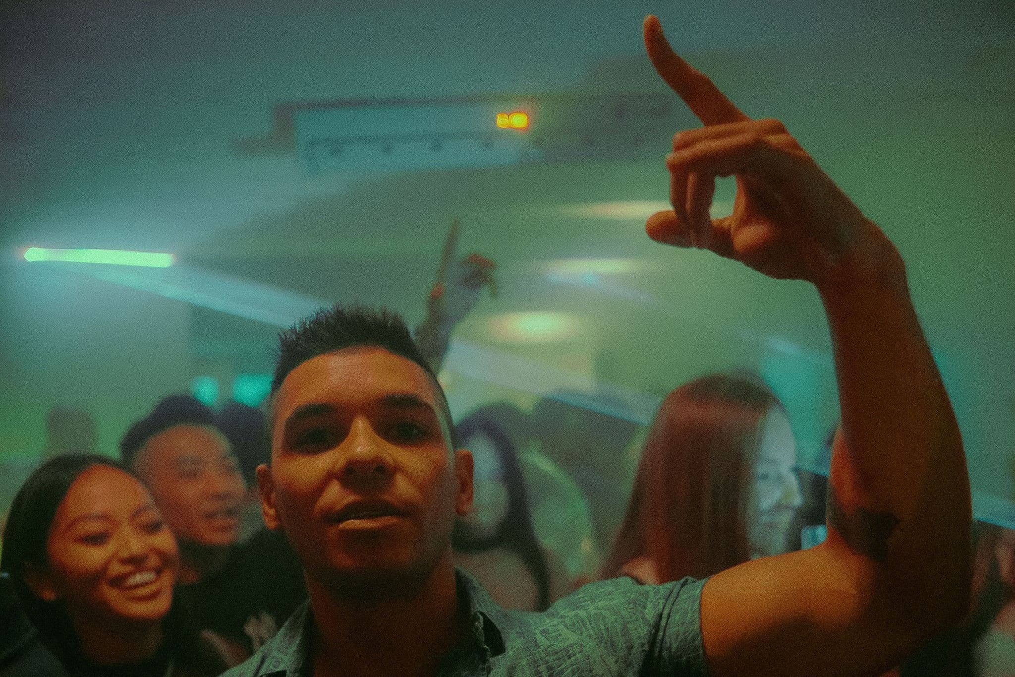 A man raises one hand on a crowded, hazy dance floor while friends smile behind him