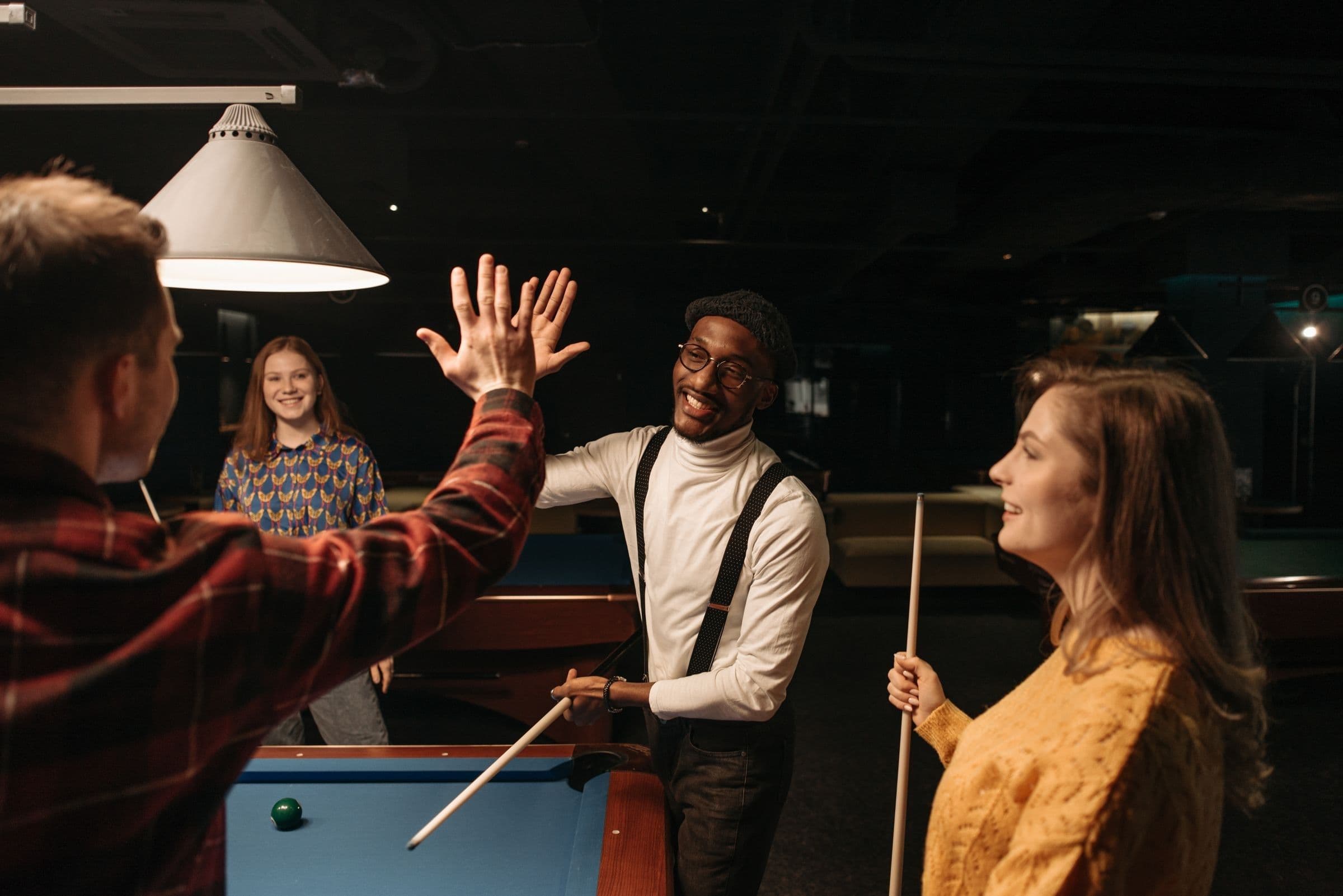 A diverse group of friends in sweaters and flannel laughing and sharing a high five at a pool hall bar after dark