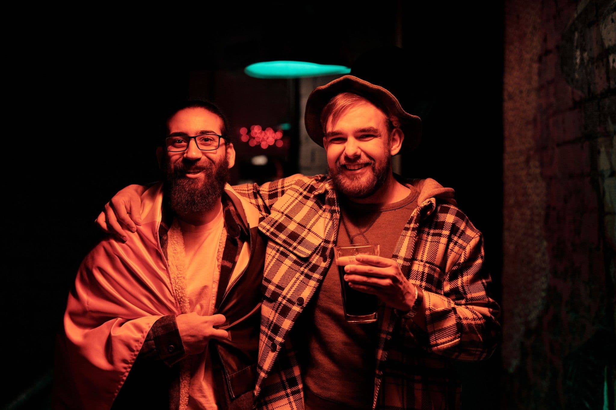 Two friends laughing arm in arm under red bar lights, one holding a drink