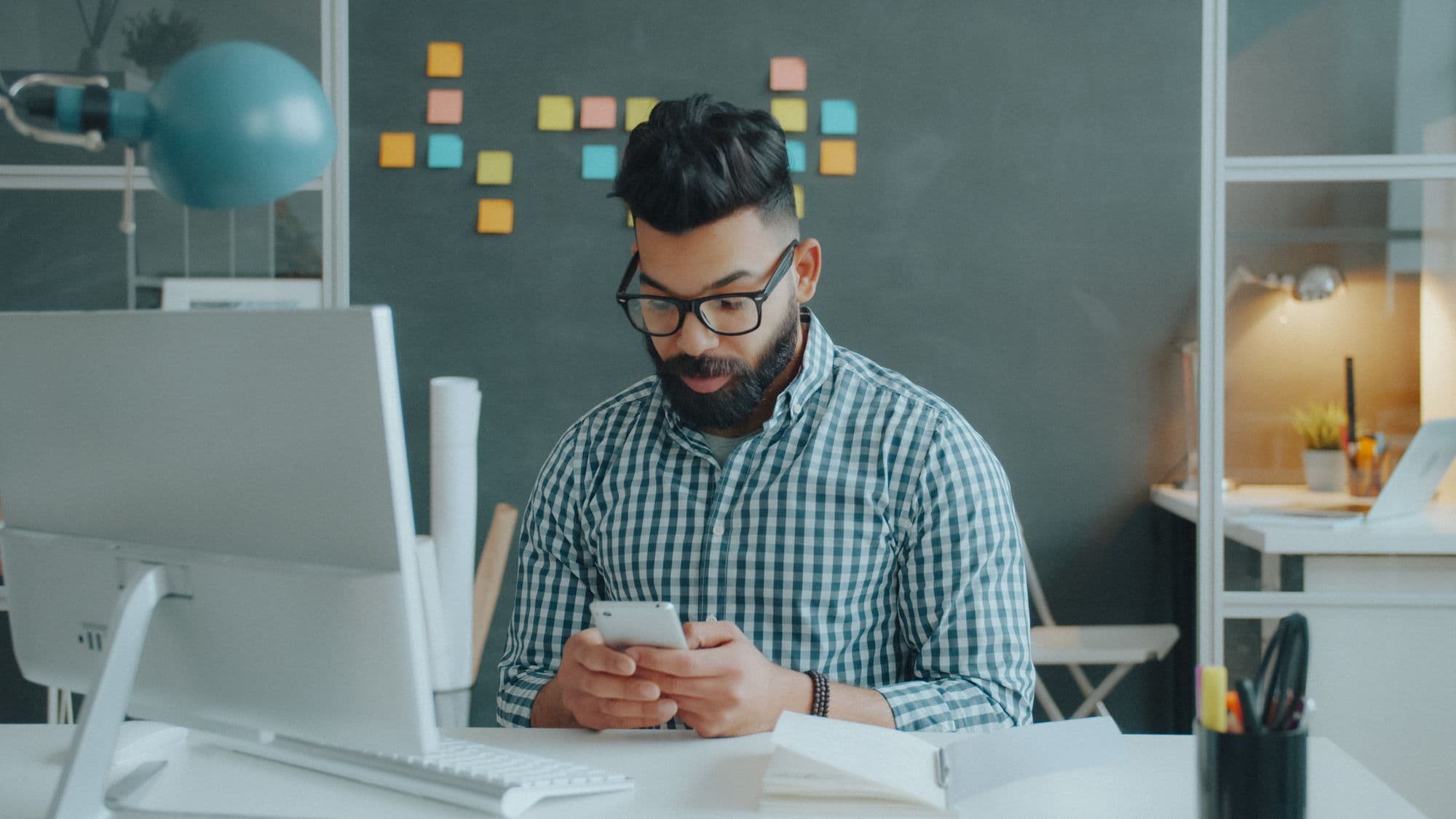 A man in glasses at a desk, looking intently at his phone